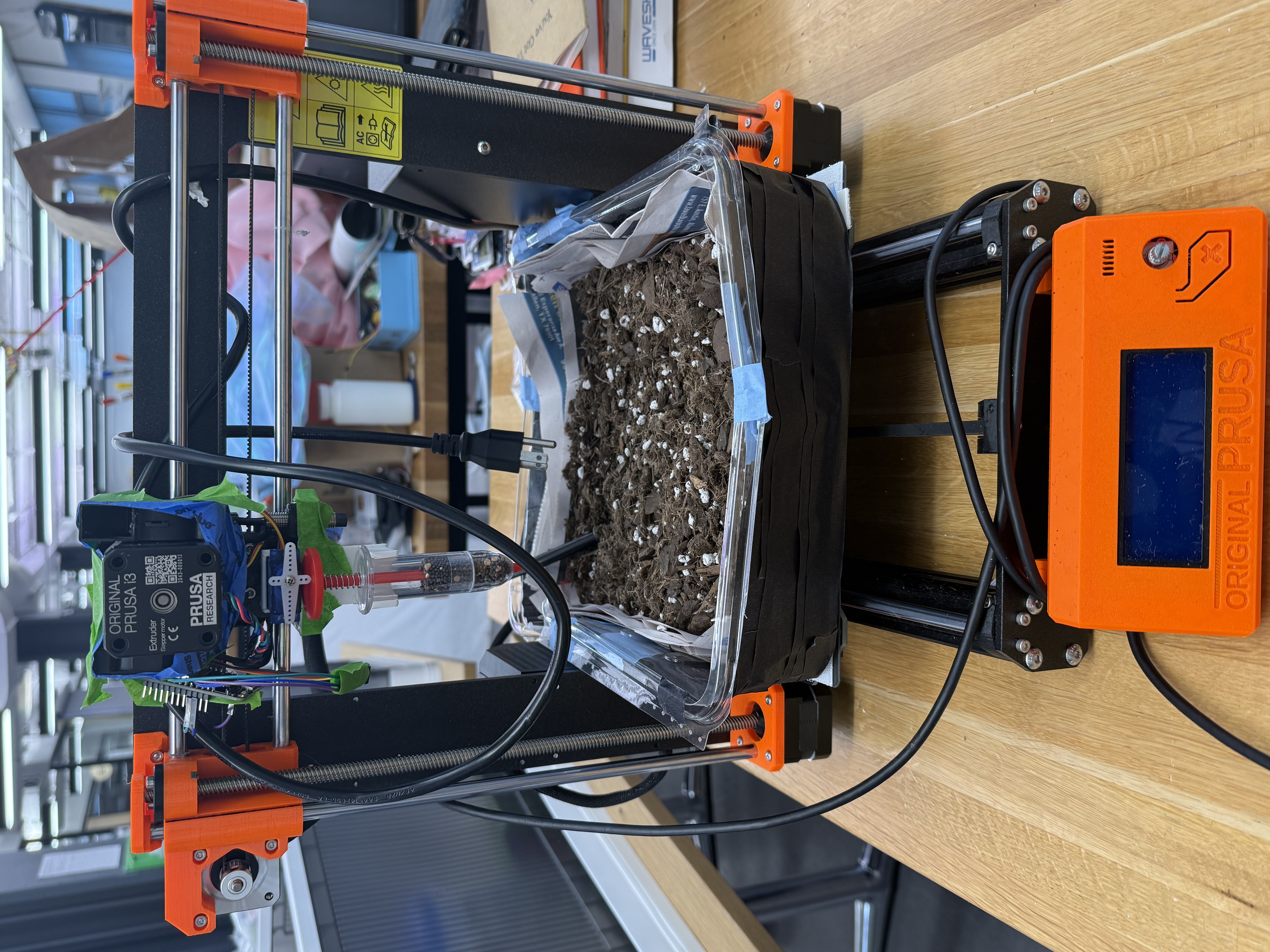 Repurposed Prusa i3 printer mounted with a seed-dropper syringe and a soil bed underneath, ready to plant